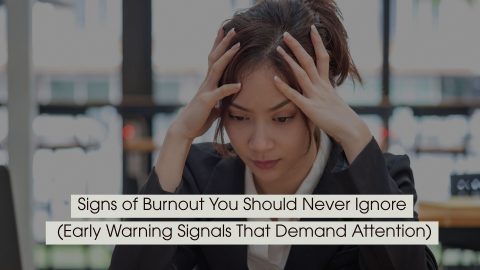 Person sitting at a desk holding their head with both hands, appearing stressed and overwhelmed, symbolizing burnout and mental fatigue, with the text “Signs of Burnout You Should Never Ignore (Early Warning Signals That Demand Attention)” displayed across the image.