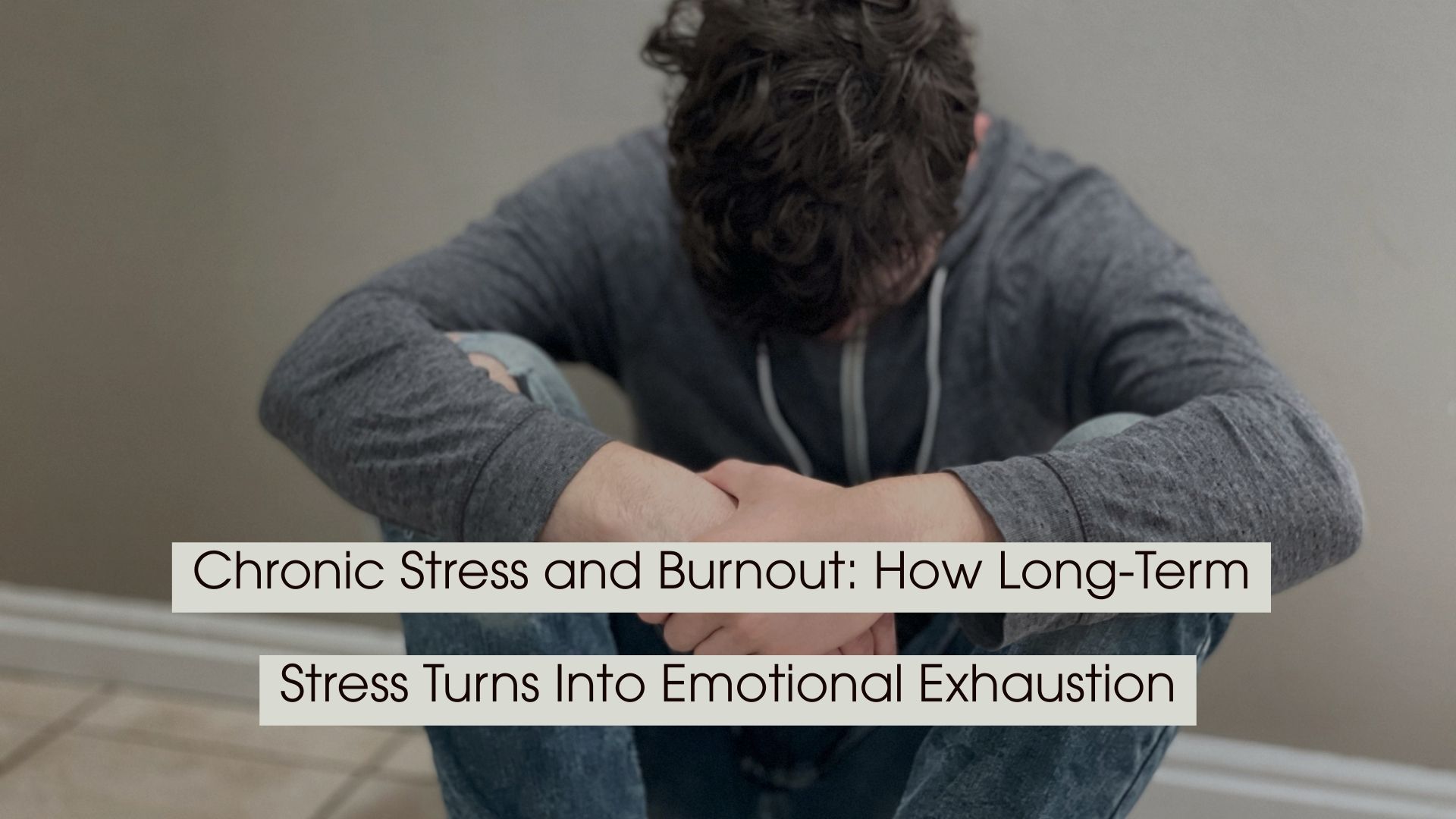 Man sitting on the floor with his head down and arms resting on his knees, appearing overwhelmed and emotionally drained, with the text “Chronic Stress and Burnout: How Long-Term Stress Turns Into Emotional Exhaustion” displayed across the image.