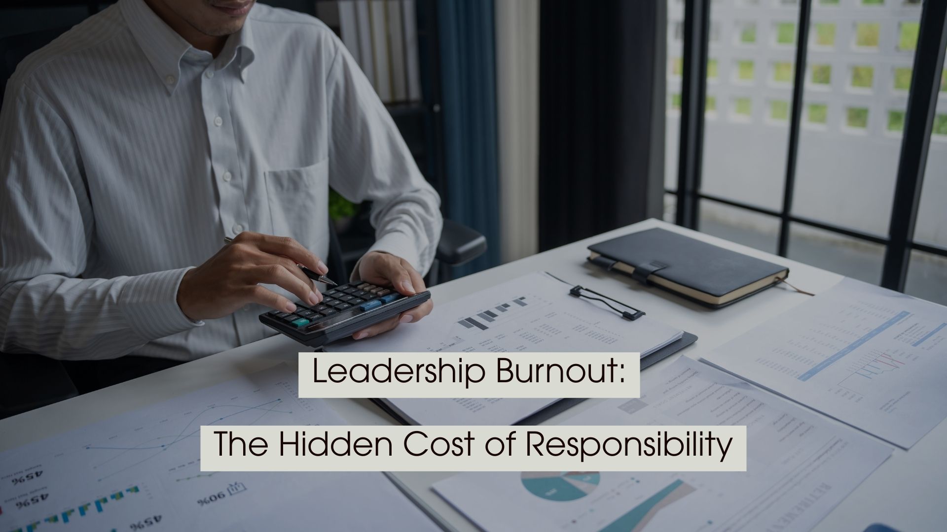 A person in a white button-up shirt sits at a desk reviewing financial documents and charts while using a calculator and holding a pen, with a notebook and paperwork spread across the workspace. The image conveys the pressure of managing responsibilities, with overlaid text reading, “Leadership Burnout: The Hidden Cost of Responsibility.”