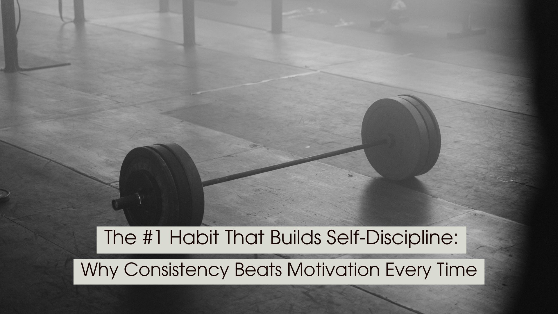 A black-and-white photo of a loaded barbell resting on the floor of an empty gym symbolizes discipline, consistency, and the quiet commitment required to achieve long-term goals. The minimalist, moody atmosphere emphasizes showing up and putting in the work even when motivation fades. Overlaid text reads: “The #1 Habit That Builds Self-Discipline: Why Consistency Beats Motivation Every Time.”