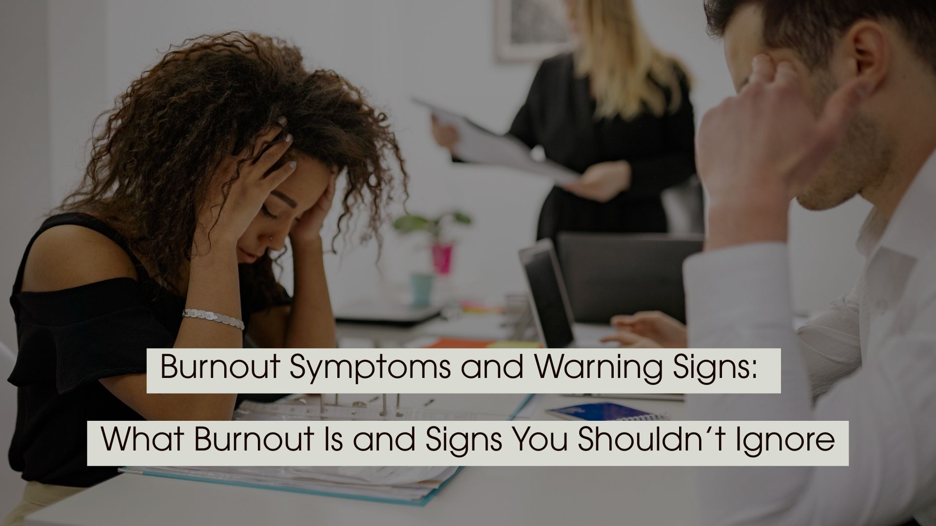 Two office workers sitting at a desk with their heads in their hands appearing stressed or exhausted, with the text “Burnout Symptoms and Warning Signs: What Burnout Is and Signs You Shouldn’t Ignore” displayed across the image.