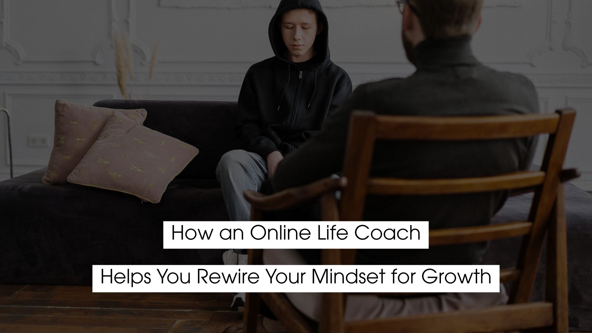 A person in a black hoodie sits on a couch with head slightly lowered, facing a life coach seated across from them in a chair, suggesting a counseling or coaching session. Overlaid text reads: ‘How an Online Life Coach Helps You Rewrite Your Mindset for Growth.’