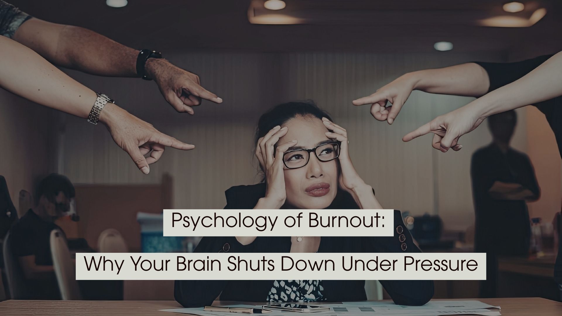 Stressed woman sitting at a desk with hands on her head while several hands point fingers at her, with the text “Psychology of Burnout: Why Your Brain Shuts Down Under Pressure” displayed across the image.