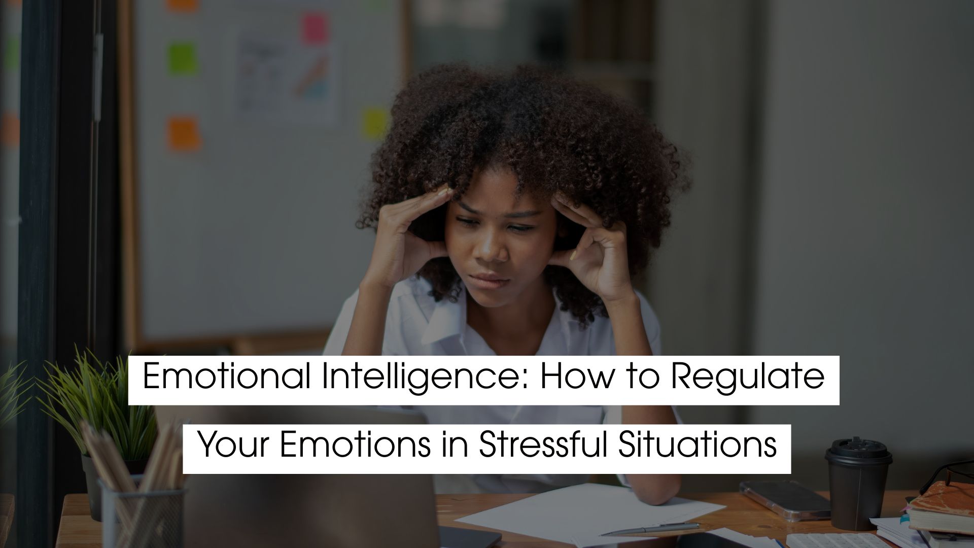 A woman sitting at a desk holds her temples while looking stressed, illustrating emotional intelligence and how to regulate emotions in stressful situations.