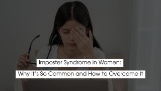 Woman sitting at a desk with eyes closed and hand on her forehead, holding glasses, appearing stressed or overwhelmed, representing imposter syndrome and self-doubt in women.