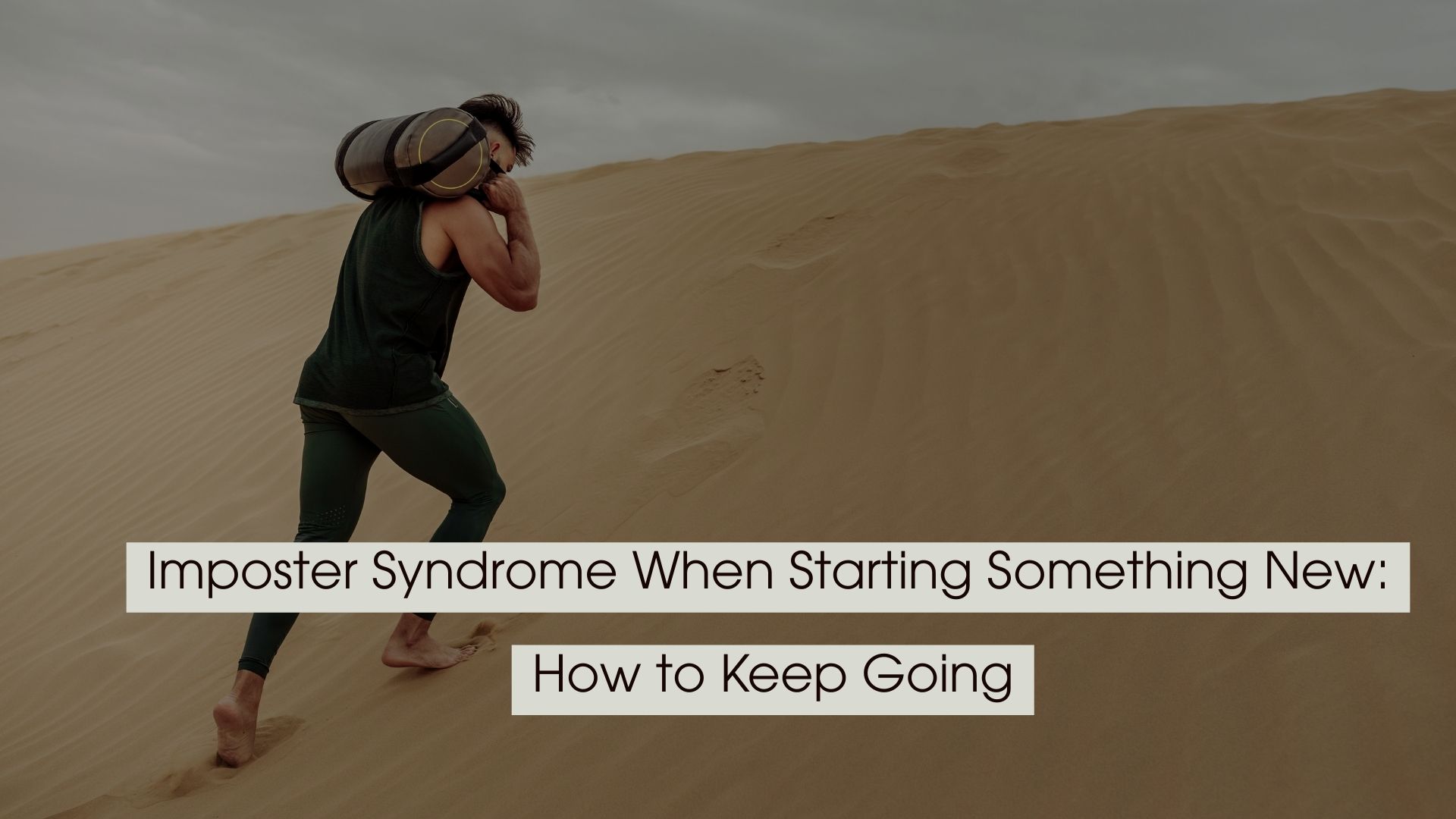 A man carrying a heavy sandbag climbs a steep sand dune under a cloudy sky, representing the challenge of overcoming imposter syndrome when starting something new.