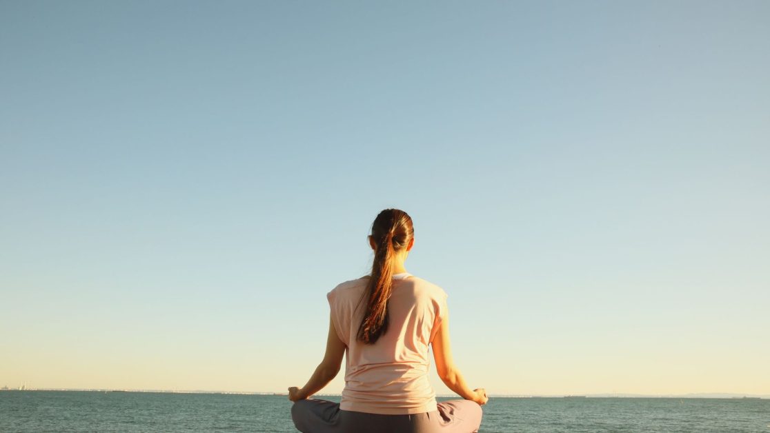A woman sitting cross-legged by the ocean, meditating peacefully under a clear sky — symbolizing the journey from overwhelm to inner peace and emotional well-being.