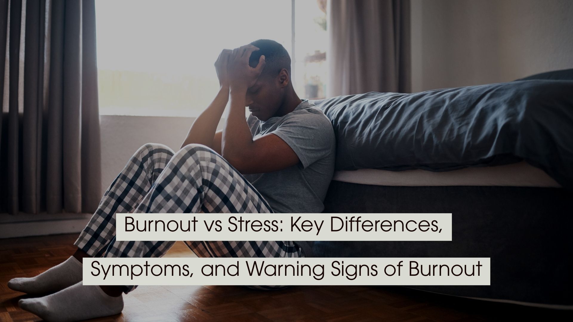 Man sitting on the floor beside a bed with his head in his hands, appearing overwhelmed or exhausted, with the text “Burnout vs Stress: Key Differences, Symptoms, and Warning Signs of Burnout” displayed across the image.